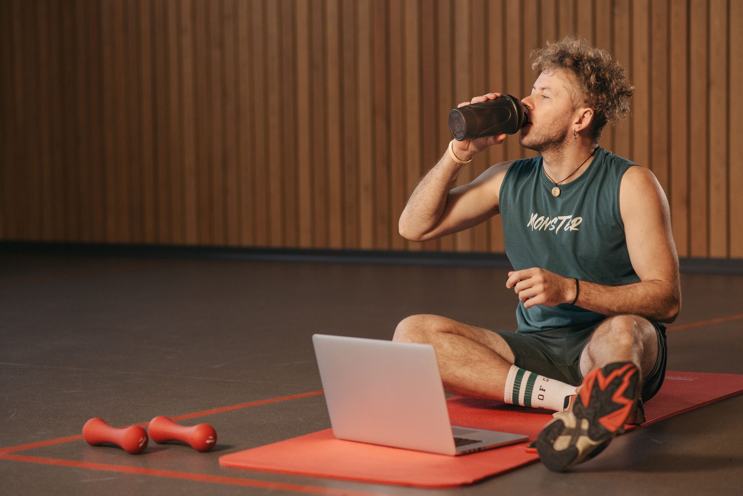 Man in gym drinking water during workout on exercise mat with laptop.